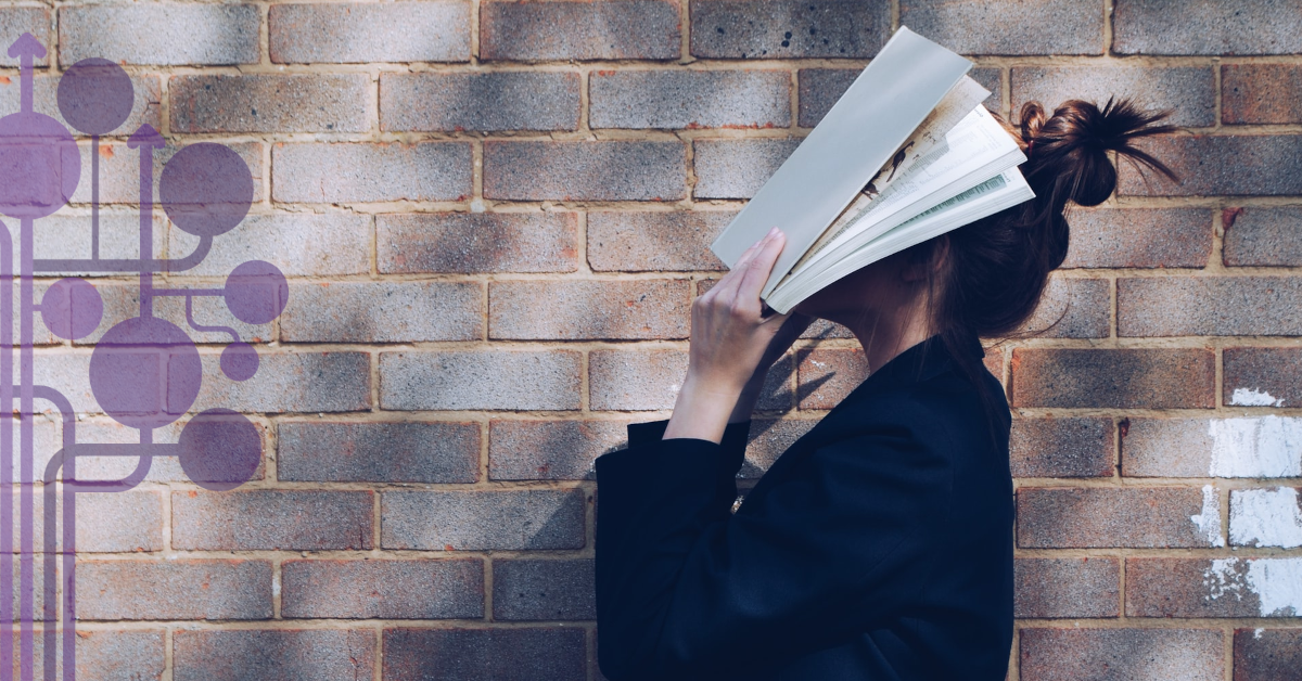 Młoda kobieta stoi bokiem na tle muru i zasłania twarz otwartą książką.