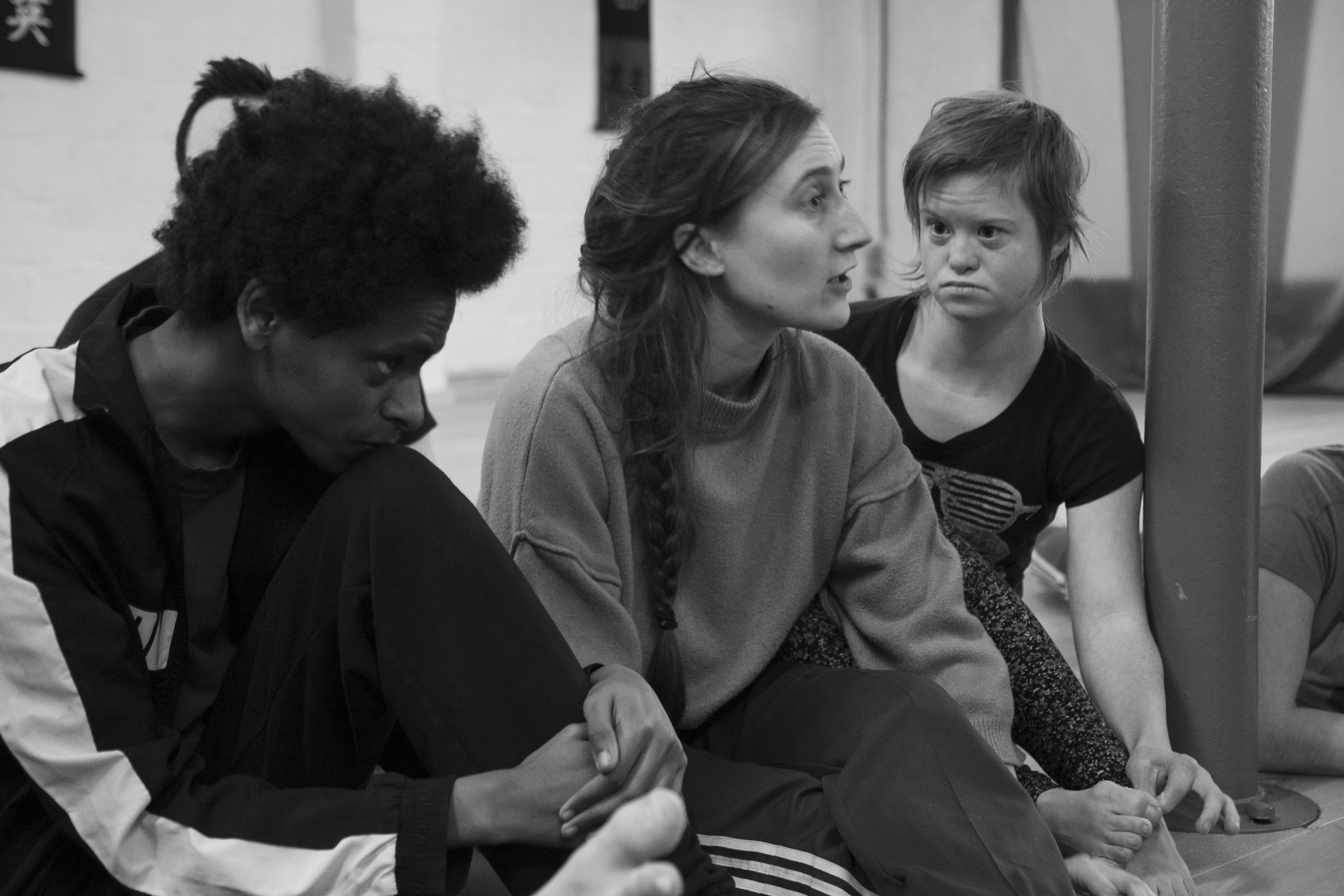 Three people sitting on a floor in a dance studio. The woman sitting in the middle is explaining something.