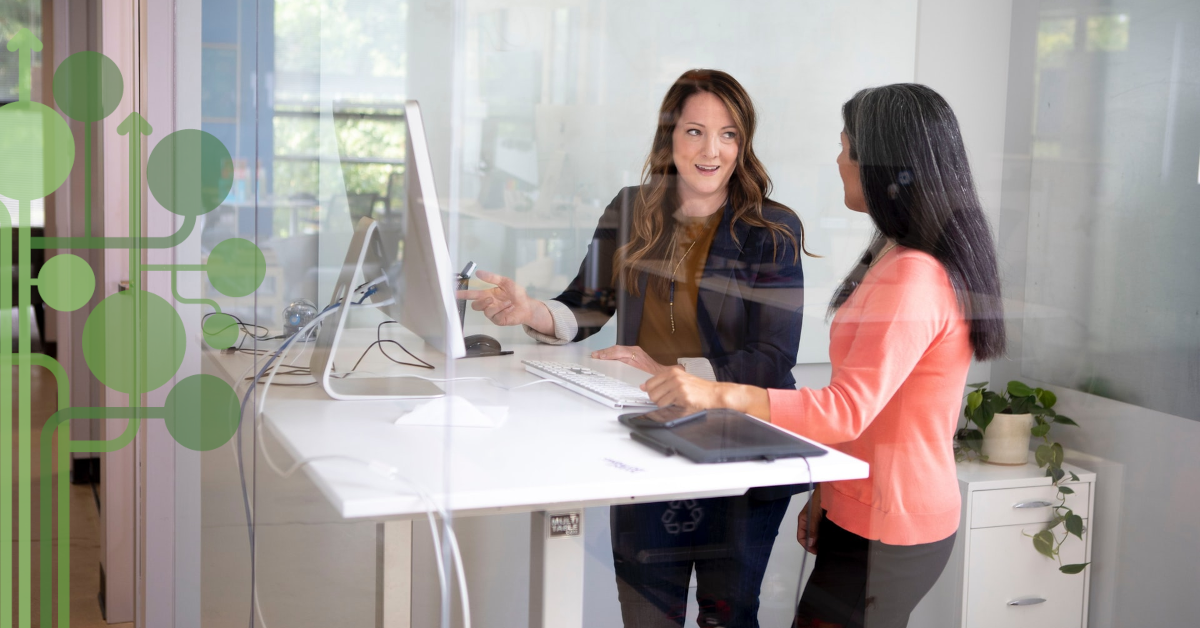 dwie kobiety stojące przy biurku z laptopem