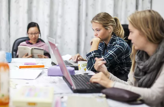 tre kvinnelige studenter sitter ved et bord og leser konsentrert, to på pc-er og en i en bok.