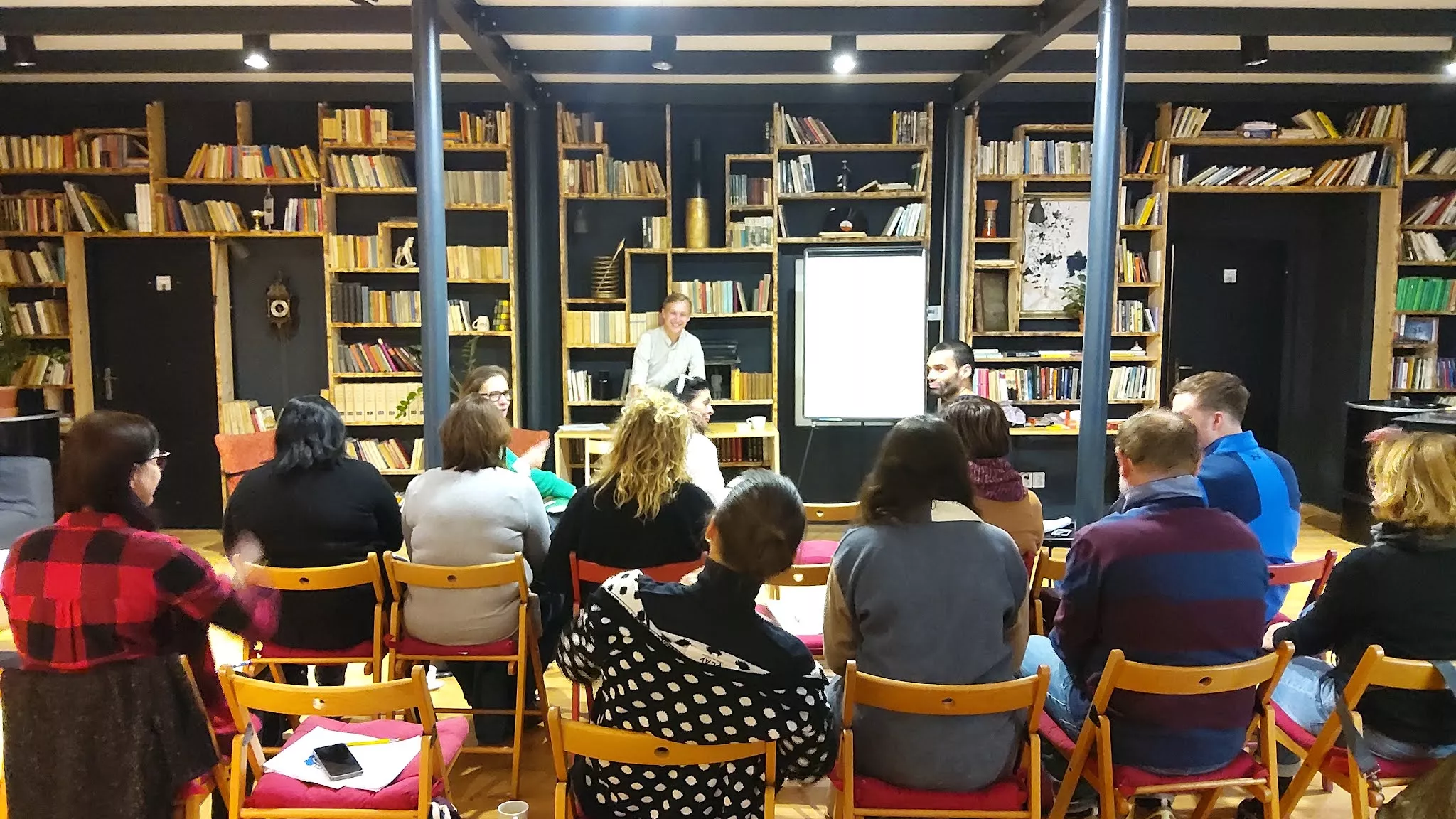 Kurs Miedzykulturowosc_Foto Urszula Maciąg Na czarnej ścianie książki. Na ich tle trenerka, a przed nią siedzący uczestnicy kursu.