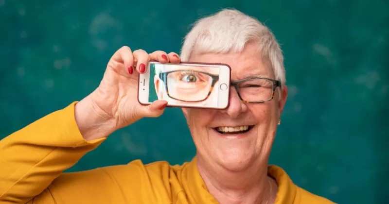 Ältere Frau mit Smartphone, in dem ihr Auge abgebildet ist, lachend.